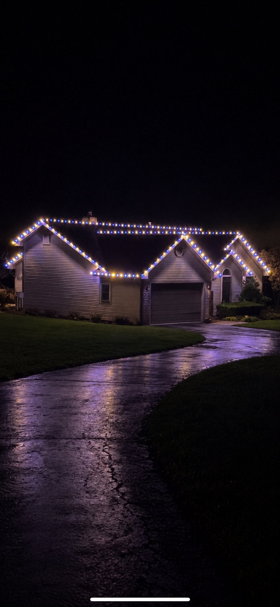 A residential house decorated with white holiday lights outlining the roofline and eaves, photographed at night with a wet driveway visible in the foreground