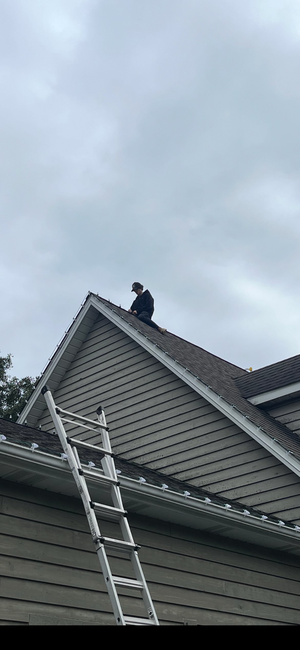 Worker sitting on pitched roof next to white ladder on residential house against cloudy sky