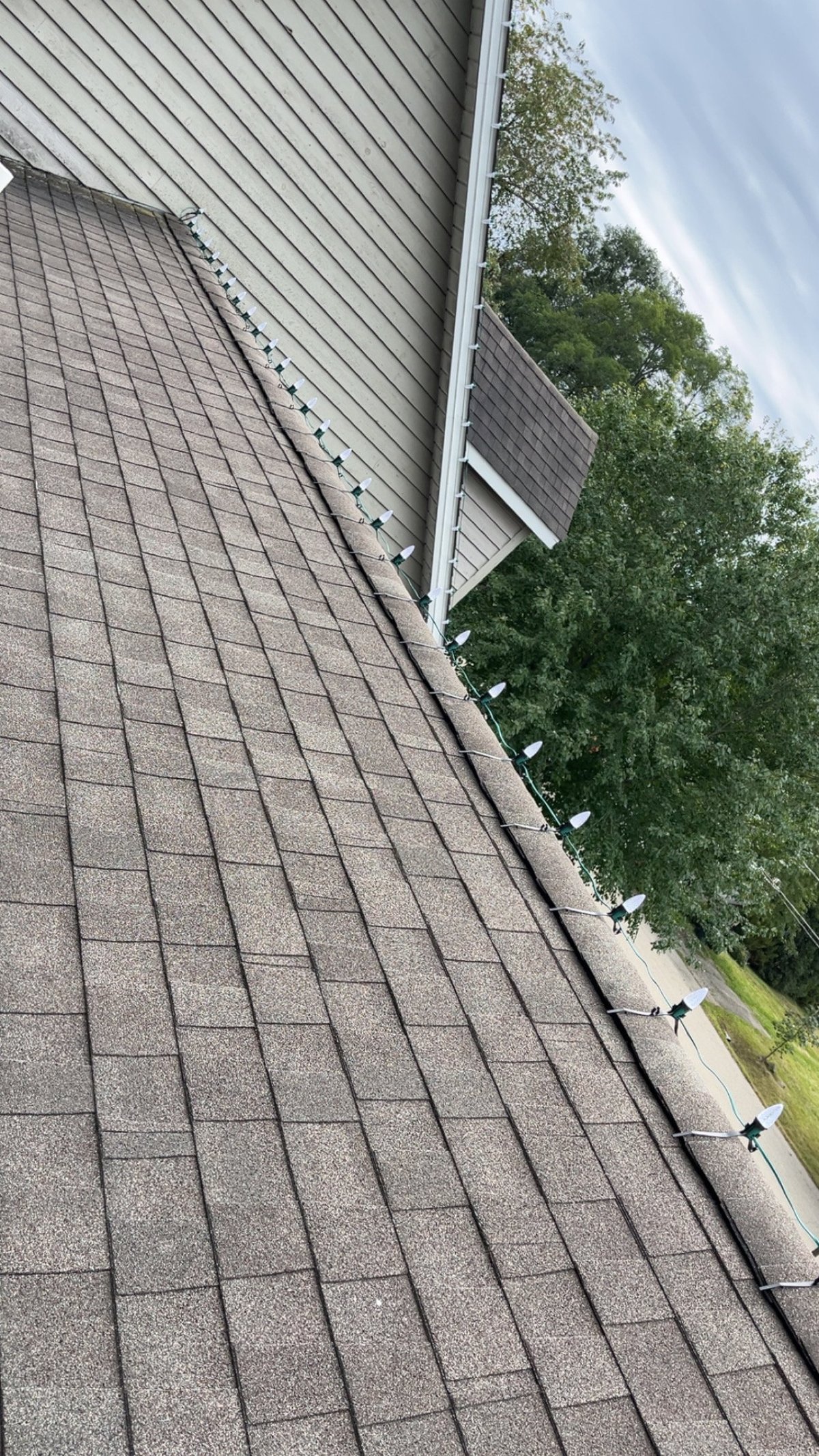 Roof with gray asphalt shingles, white gutters, and metal flashing against a cloudy sky with green trees visible
