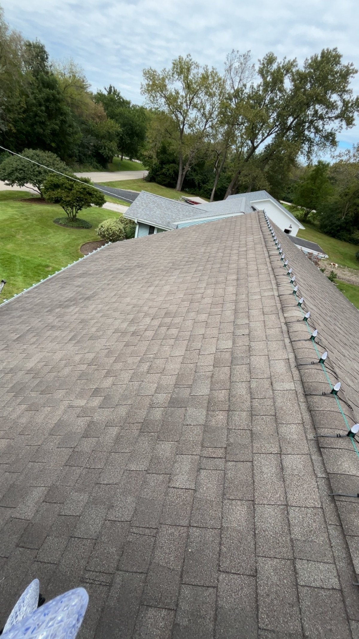 Roofing contractor standing on sloped asphalt shingle roof of modern house with landscaped lawn and trees in background
