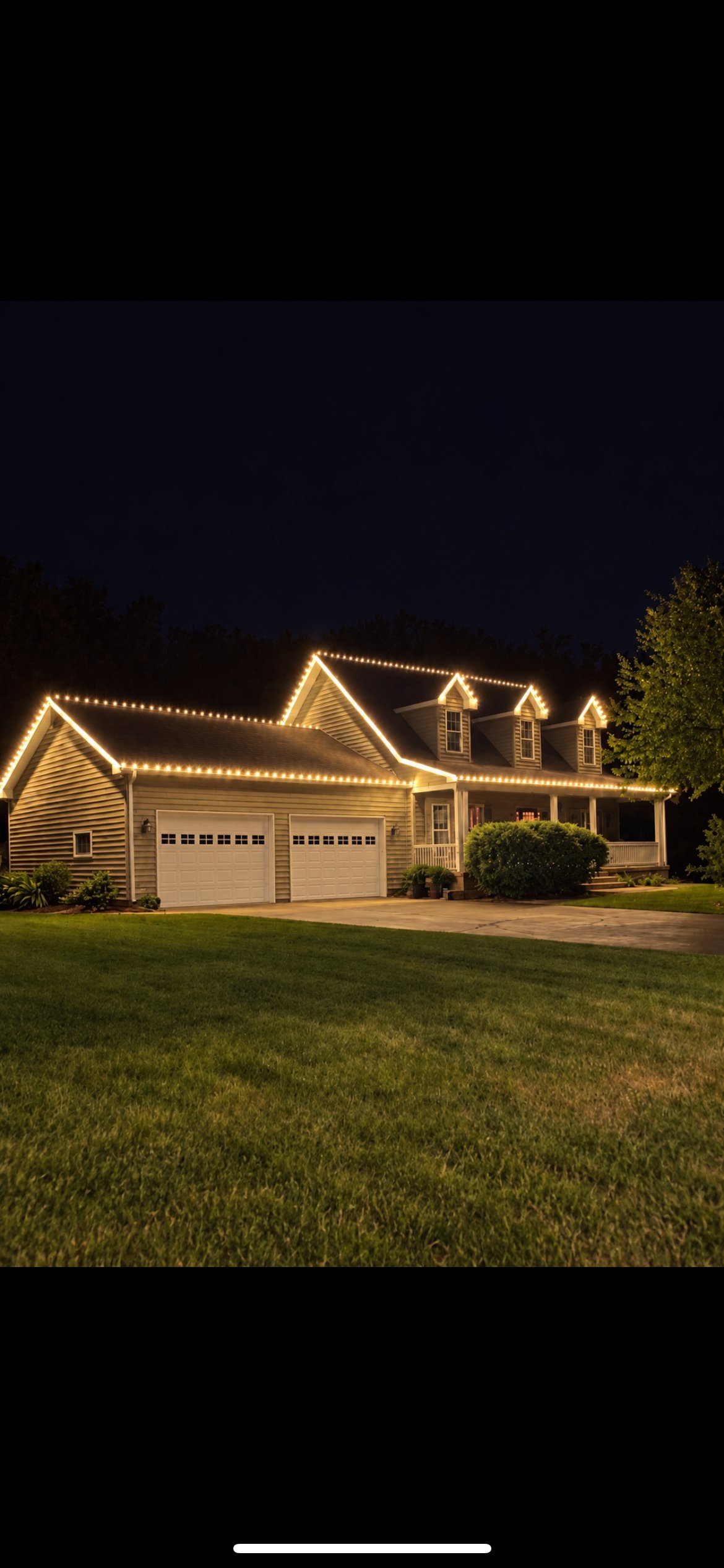 House at night decorated with white Christmas lights along the roofline and gables, with wet driveway visible in foreground